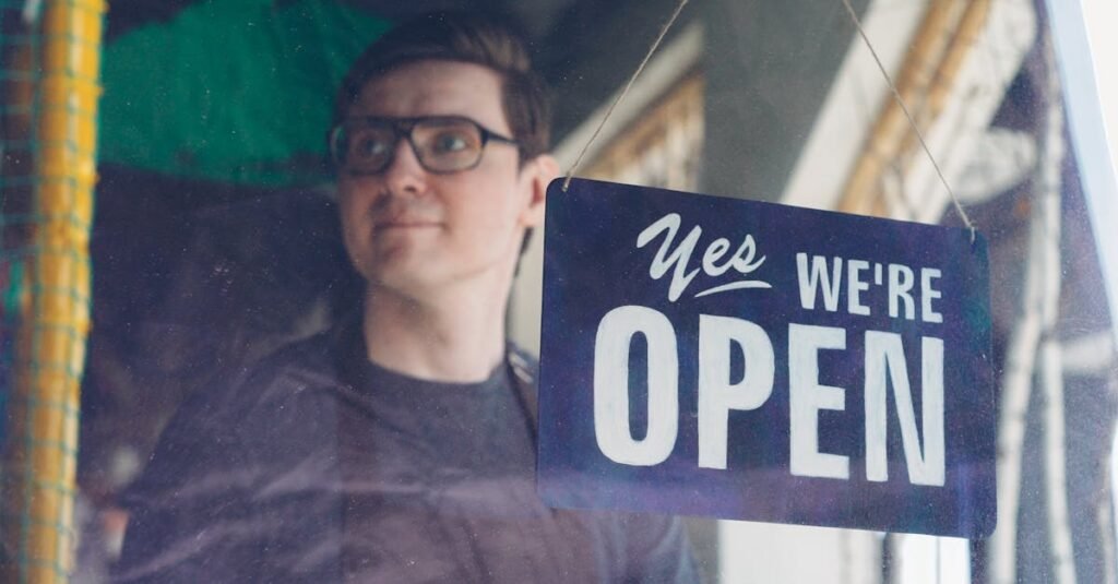 Smiling adult man behind a glass with a 'Yes, We're Open' sign, suggesting a welcoming store.