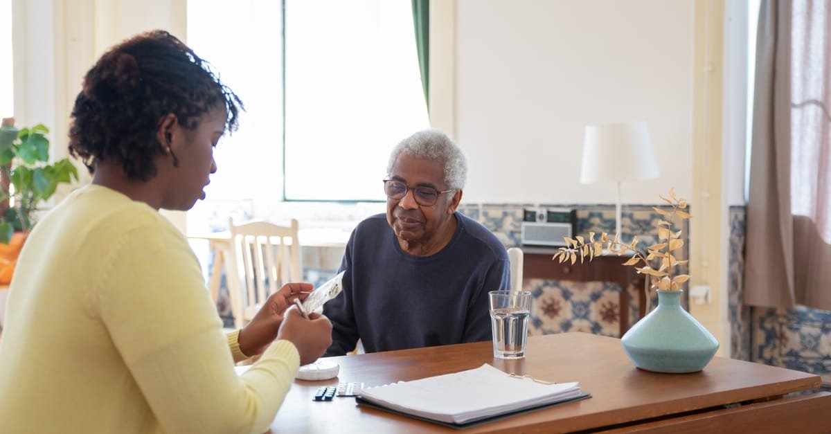 A caregiver assists an elderly man with medication in a warm, sunlit living room.